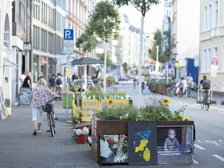 Begrünte Stadtstraße mit Fahrradspur, Hochbeeten, Kunstmotiven und Menschen zu Fuß und mit dem Rad.