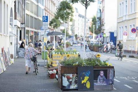 Begrünte Stadtstraße mit Fahrradspur, Hochbeeten, Kunstmotiven und Menschen zu Fuß und mit dem Rad.