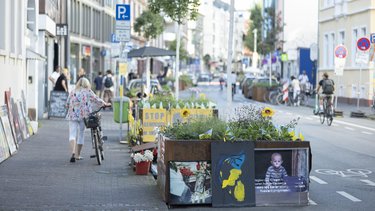 Begrünte Stadtstraße mit Fahrradspur, Hochbeeten, Kunstmotiven und Menschen zu Fuß und mit dem Rad.