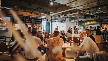 Gruppen von Menschen sitzen an langen Holztischen in einer offenen Gastronomiefläche.