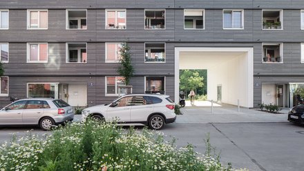 Straßenansicht eines Wohngebäudes mit parkenden Autos und offenem Durchgang zum Innenhof.