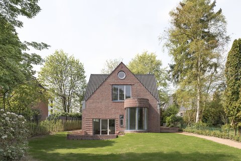 Einfamilienhaus aus rotem Backstein mit Satteldach, großer Rasenfläche und Bäumen im Garten.