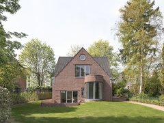 Einfamilienhaus aus rotem Backstein mit Satteldach, großer Rasenfläche und Bäumen im Garten.