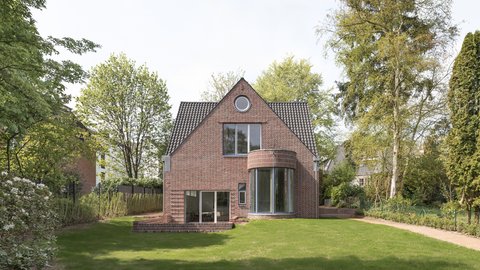 Einfamilienhaus aus rotem Backstein mit Satteldach, großer Rasenfläche und Bäumen im Garten.
