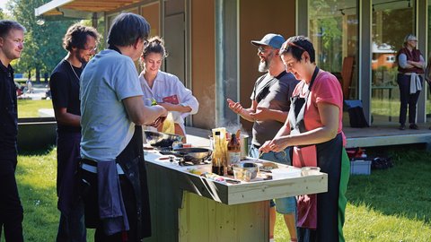 "Eine Gruppe von Personen beim gemeinsamen Kochen im Freien. Eine Frau mit Schürze bereitet Speisen an einer provisorischen Holzküche zu, während neben ihr zwei Männer miteinander sprechen. Im Hintergrund steht ein modernes Holzgebäude mit großen Fenstern."