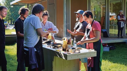 "Eine Gruppe von Personen beim gemeinsamen Kochen im Freien. Eine Frau mit Schürze bereitet Speisen an einer provisorischen Holzküche zu, während neben ihr zwei Männer miteinander sprechen. Im Hintergrund steht ein modernes Holzgebäude mit großen Fenstern."