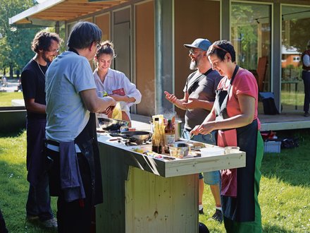 "Eine Gruppe von Personen beim gemeinsamen Kochen im Freien. Eine Frau mit Schürze bereitet Speisen an einer provisorischen Holzküche zu, während neben ihr zwei Männer miteinander sprechen. Im Hintergrund steht ein modernes Holzgebäude mit großen Fenstern."
