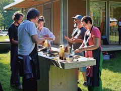 "Eine Gruppe von Personen beim gemeinsamen Kochen im Freien. Eine Frau mit Schürze bereitet Speisen an einer provisorischen Holzküche zu, während neben ihr zwei Männer miteinander sprechen. Im Hintergrund steht ein modernes Holzgebäude mit großen Fenstern."