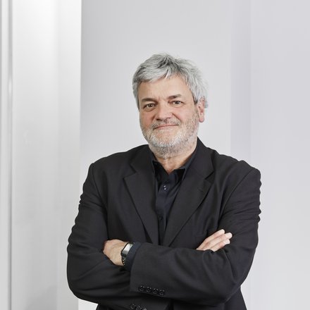 Ein Porträt zeigt eine einzelne erwachsene Person mit grauem Haar und Bart in dunkler Kleidung vor hellem, neutralem Hintergrund, die Arme sind verschränkt und der Blick ist ruhig auf die Kamera gerichtet, wodurch das Bild eine professionelle und souveräne Darstellung vermittelt und für fachliche Autorität, Erfahrung und persönliche Verantwortung innerhalb eines architektonischen oder kulturellen Kontextes steht.