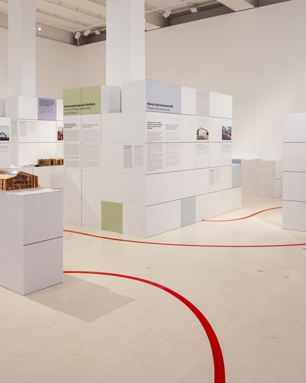 Helle Ausstellung mit weißen, würfelförmigen Stellwänden voller Text‑ und Bildtafeln in Pastellfarben; roter Orientierungsstreifen auf dem Boden; vorn ein Holz‑Architekturmodell auf Podest; rechts ein Monitor.