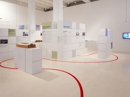 Helle Ausstellung mit weißen, würfelförmigen Stellwänden voller Text‑ und Bildtafeln in Pastellfarben; roter Orientierungsstreifen auf dem Boden; vorn ein Holz‑Architekturmodell auf Podest; rechts ein Monitor.