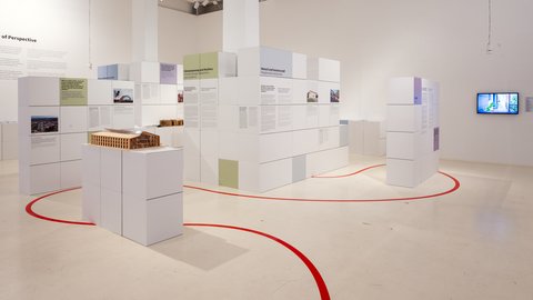 Helle Ausstellung mit weißen, würfelförmigen Stellwänden voller Text‑ und Bildtafeln in Pastellfarben; roter Orientierungsstreifen auf dem Boden; vorn ein Holz‑Architekturmodell auf Podest; rechts ein Monitor.