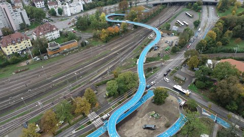 Luftaufnahme einer geschwungenen blauen Radbrücke, die Straßen und Bahntrassen überquert.