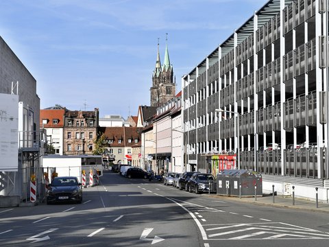 Straßenansicht einer europäischen Stadt mit gotischem Kirchturm mit grünen und goldenen Spitzen in der Bildmitte. Links moderne graue Gebäude, rechts mehrgeschossige weiße Fassade. Asphaltstraße mit Fahrspuren, geparkten Autos und Baustellenabsperrung. Blauer Himmel mit leichten Wolken.