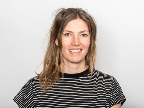 Lächelnde Person mit schulterlangem braunem Haar in schwarz-weiß gestreiftem T-Shirt vor hellem Hintergrund.