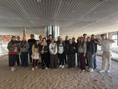 „Gruppenfoto der Exkursionsteilnehmer in einer weitläufigen Halle mit industrieller Gitterdecke und beigem Bodenbelag.“