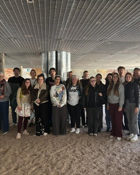 „Gruppenfoto der Exkursionsteilnehmer in einer weitläufigen Halle mit industrieller Gitterdecke und beigem Bodenbelag.“