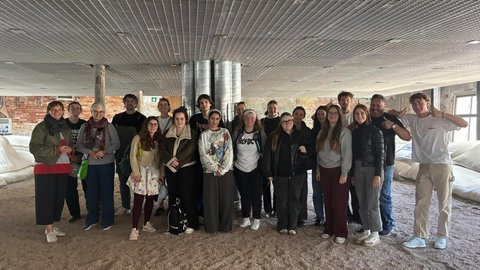 „Gruppenfoto der Exkursionsteilnehmer in einer weitläufigen Halle mit industrieller Gitterdecke und beigem Bodenbelag.“