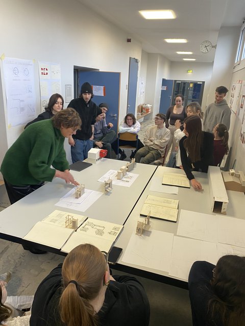 „Eine Dozentin im grünen Pullover bespricht Architekturmodelle aus Holz und Skizzen mit einer Gruppe von Studierenden an einem Tisch.“