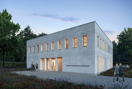 Außenansicht: Zweigeschossiger, heller Mauerwerksbau in Dämmerung; schmale Fenster mit warmem Licht, gepflasterter Vorplatz und Wildblumen; an der Fassade steht „Verband für ländliche Entwicklung“.