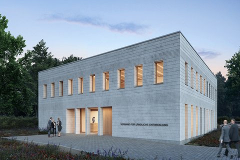 Außenansicht: Zweigeschossiger, heller Mauerwerksbau in Dämmerung; schmale Fenster mit warmem Licht, gepflasterter Vorplatz und Wildblumen; an der Fassade steht „Verband für ländliche Entwicklung“.