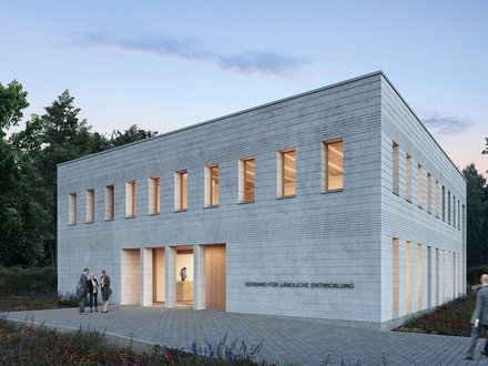 Außenansicht: Zweigeschossiger, heller Mauerwerksbau in Dämmerung; schmale Fenster mit warmem Licht, gepflasterter Vorplatz und Wildblumen; an der Fassade steht „Verband für ländliche Entwicklung“.