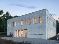 Außenansicht: Zweigeschossiger, heller Mauerwerksbau in Dämmerung; schmale Fenster mit warmem Licht, gepflasterter Vorplatz und Wildblumen; an der Fassade steht „Verband für ländliche Entwicklung“.