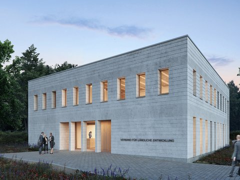 Außenansicht: Zweigeschossiger, heller Mauerwerksbau in Dämmerung; schmale Fenster mit warmem Licht, gepflasterter Vorplatz und Wildblumen; an der Fassade steht „Verband für ländliche Entwicklung“.