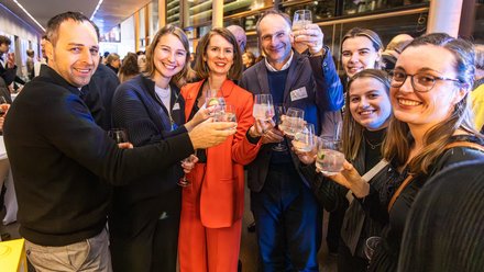 Eine Gruppe von Menschen steht eng zusammen, hält Gläser in die Höhe und lächelt in die Kamera, im Hintergrund ist eine größere Veranstaltungssituation erkennbar, wodurch das Bild einen gemeinsamen Moment des Feierns festhält und für Gemeinschaft, Anerkennung und das soziale Miteinander im Rahmen einer kulturellen oder fachlichen Veranstaltung steht.