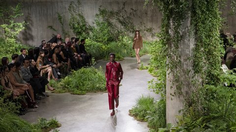 "Ein Model in einem roten Lederoutfit läuft über einen geschwungenen Laufsteg durch eine dichte Pflanzeninstallation in einer Betonhalle. Das Publikum sitzt dicht am Rand und beobachtet die Präsentation. Im Hintergrund folgt ein weiteres Model in einem schlichten Outfit. Kletterpflanzen wachsen an den Wänden und Säulen, während Spotlights die Szene ausleuchten."