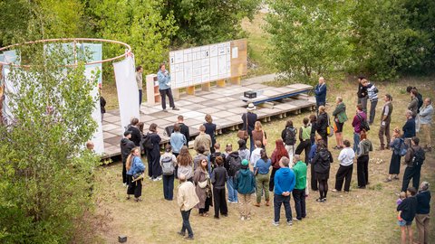 „Eine Menschenmenge steht im Freien auf einer Wiese und lauscht einem Redner, der auf einer flachen Holzplattform mit aufgestellten weißen Infotafeln steht.“