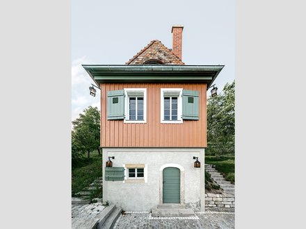 Ein kleines, freistehendes Haus mit rechteckigem Grundriss steht am Hang in einer grünen Umgebung, der untere Teil ist verputzt und massiv, der obere Teil besteht aus vertikal verschalten Holzflächen in warmem Farbton, grüne Fensterläden, ein steiles Ziegeldach und ein gemauerter Schornstein prägen das Erscheinungsbild, eine Außentreppe führt seitlich nach oben, wodurch das Bild ein sorgfältig instand gesetztes oder neu interpretiertes Weinberg- oder Gartenhaus zeigt, das traditionelle Bauweise mit zeitgemäßer Nutzung verbindet und für den behutsamen Umgang mit kleinteiliger historischer Architektur und Landschaft steht.