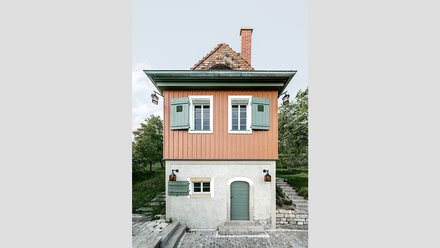 Ein kleines, freistehendes Haus mit rechteckigem Grundriss steht am Hang in einer grünen Umgebung, der untere Teil ist verputzt und massiv, der obere Teil besteht aus vertikal verschalten Holzflächen in warmem Farbton, grüne Fensterläden, ein steiles Ziegeldach und ein gemauerter Schornstein prägen das Erscheinungsbild, eine Außentreppe führt seitlich nach oben, wodurch das Bild ein sorgfältig instand gesetztes oder neu interpretiertes Weinberg- oder Gartenhaus zeigt, das traditionelle Bauweise mit zeitgemäßer Nutzung verbindet und für den behutsamen Umgang mit kleinteiliger historischer Architektur und Landschaft steht.