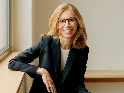 Porträt einer Frau mittleren Alters mit schulterlangem, honigblondem Haar und rechteckiger Brille mit dunklem Rahmen. Sie lächelt freundlich in die Kamera. Sie trägt einen schwarzen Blazer über einem hellen Shirt. Die Frau sitzt entspannt auf einer hellen Fensterbank, ihr linker Arm ruht auf ihrem Oberschenkel. Im Hintergrund helle, neutrale Wand und ein Fenster mit grauem Rahmen. Die Beleuchtung ist natürlich und warm.