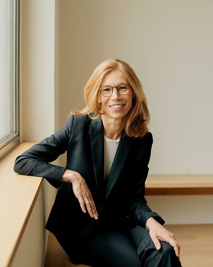 Porträt einer Frau mittleren Alters mit schulterlangem, honigblondem Haar und rechteckiger Brille mit dunklem Rahmen. Sie lächelt freundlich in die Kamera. Sie trägt einen schwarzen Blazer über einem hellen Shirt. Die Frau sitzt entspannt auf einer hellen Fensterbank, ihr linker Arm ruht auf ihrem Oberschenkel. Im Hintergrund helle, neutrale Wand und ein Fenster mit grauem Rahmen. Die Beleuchtung ist natürlich und warm.