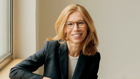 Porträt einer Frau mittleren Alters mit schulterlangem, honigblondem Haar und rechteckiger Brille mit dunklem Rahmen. Sie lächelt freundlich in die Kamera. Sie trägt einen schwarzen Blazer über einem hellen Shirt. Die Frau sitzt entspannt auf einer hellen Fensterbank, ihr linker Arm ruht auf ihrem Oberschenkel. Im Hintergrund helle, neutrale Wand und ein Fenster mit grauem Rahmen. Die Beleuchtung ist natürlich und warm.