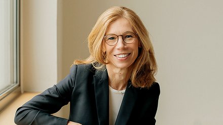 Porträt einer Frau mittleren Alters mit schulterlangem, honigblondem Haar und rechteckiger Brille mit dunklem Rahmen. Sie lächelt freundlich in die Kamera. Sie trägt einen schwarzen Blazer über einem hellen Shirt. Die Frau sitzt entspannt auf einer hellen Fensterbank, ihr linker Arm ruht auf ihrem Oberschenkel. Im Hintergrund helle, neutrale Wand und ein Fenster mit grauem Rahmen. Die Beleuchtung ist natürlich und warm.