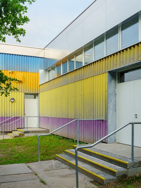 Detailansicht der farbenfrohen Wellblechfassade des Neubaus. Die Streifen sind lila, gelb und blau. Oben ist der Schriftzug "CRAZY" in großen, unregelmäßigen, lilafarbenen Buchstaben angebracht. Darunter befindet sich ein Fenster. Ein grüner, runder Lautsprecher ist links an der gelben Wand befestigt.