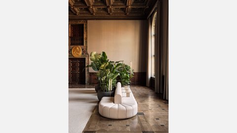 Ein heller Loungebereich mit modernem Sofa und großen Zimmerpflanzen in einem historischen Saal mit hoher Kassettendecke und großen Fenstern.