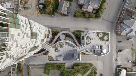 Draufsicht auf die Dachterrassen eines kompakten Gebäudeensembles: geschwungene Holzdecks, Sitzbereiche und Pflanzflächen bilden zusammenhängende Außenräume, links daran schließt ein hoher Turm mit gestaffelten Fenstern an; umliegende Straßen und Gebäude sind aus der Vogelperspektive sichtbar.