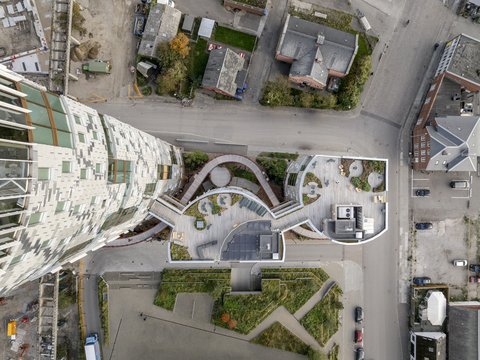 Draufsicht auf die Dachterrassen eines kompakten Gebäudeensembles: geschwungene Holzdecks, Sitzbereiche und Pflanzflächen bilden zusammenhängende Außenräume, links daran schließt ein hoher Turm mit gestaffelten Fenstern an; umliegende Straßen und Gebäude sind aus der Vogelperspektive sichtbar.