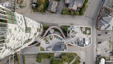 Draufsicht auf die Dachterrassen eines kompakten Gebäudeensembles: geschwungene Holzdecks, Sitzbereiche und Pflanzflächen bilden zusammenhängende Außenräume, links daran schließt ein hoher Turm mit gestaffelten Fenstern an; umliegende Straßen und Gebäude sind aus der Vogelperspektive sichtbar.
