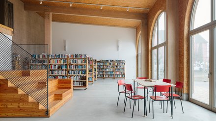  Helle Bibliothek mit hohen Rundbogenfenstern; mobile Bücherregale an einer weißen Wand, links eine Holztreppe mit Sitzstufen, vorn ein Tisch mit roten Stühlen.