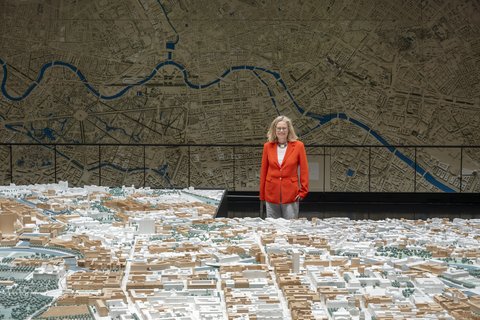 „Eine Frau im roten Blazer betrachtet ein großes, detailliertes 3D-Stadtmodell vor einer raumhohen Wandkarte mit Flussverlauf.“