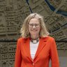 „Eine Frau im roten Blazer betrachtet ein großes, detailliertes 3D-Stadtmodell vor einer raumhohen Wandkarte mit Flussverlauf.“