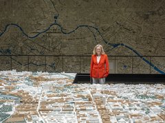 „Eine Frau im roten Blazer betrachtet ein großes, detailliertes 3D-Stadtmodell vor einer raumhohen Wandkarte mit Flussverlauf.“