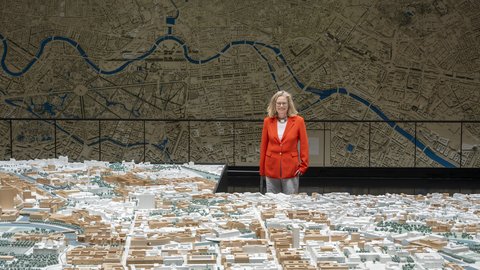 „Eine Frau im roten Blazer betrachtet ein großes, detailliertes 3D-Stadtmodell vor einer raumhohen Wandkarte mit Flussverlauf.“