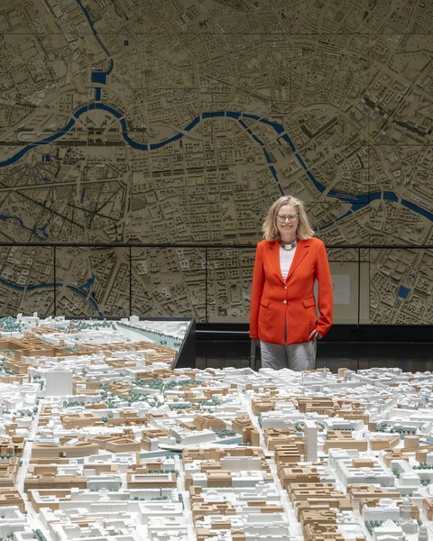 „Eine Frau im roten Blazer betrachtet ein großes, detailliertes 3D-Stadtmodell vor einer raumhohen Wandkarte mit Flussverlauf.“