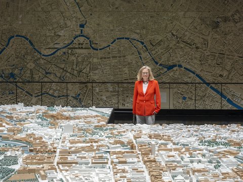 „Eine Frau im roten Blazer betrachtet ein großes, detailliertes 3D-Stadtmodell vor einer raumhohen Wandkarte mit Flussverlauf.“