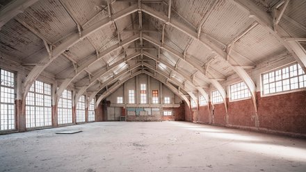 Große leerstehende Industriehalle mit hohen Fenstern, Backsteinwänden und sichtbarer Holzdachkonstruktion.